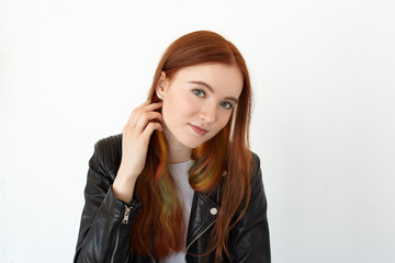 Horizontal portrait of charming red-haired student female with green eyes in a black leather jacket looking thoughtful and dreamy at camera touching her hair resting between classes at the University