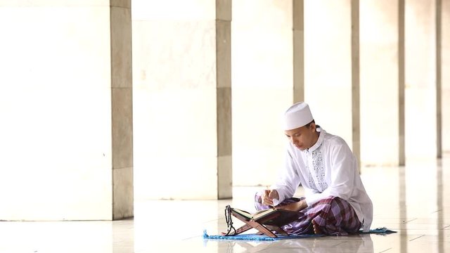 Young religious muslim man reading Quran in the mosque while sitting and wearing islamic clothes