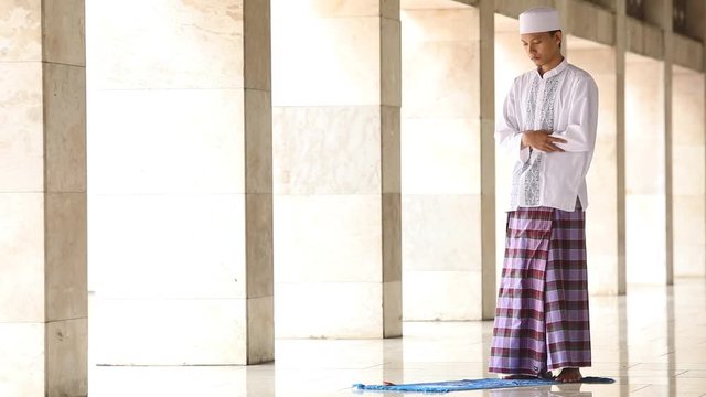  
Young muslim person wearing islamic clothes and praying in the mosque with poses: standing, raise hands, and bowing
