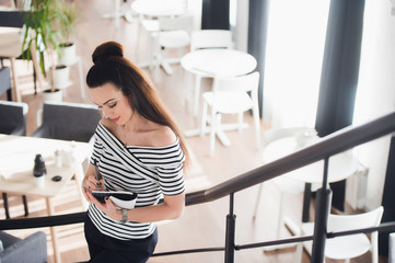 Photo of pretty woman making notes and thinking over a subject while standing in some coffee shop with her laptop near big window.