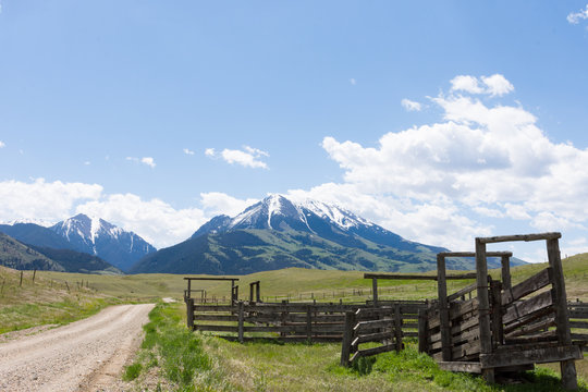 An old wooden corral in Paradise Valley Montana next to a gravel road that leads to the rugged, snow capped mountains in the distance. Fluffy white clouds above.