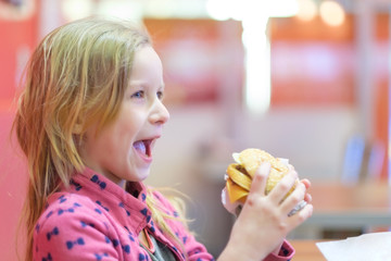 Adorable blonde cheerful girl in pink clothes eat double burger with beef and cheese in fast food restaurant
