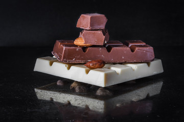 Pieces of milk chocolate with almonds and tiles of white chocolate with hazelnuts on a dark old glossy background