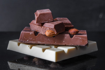 Pieces of milk chocolate with almonds and tiles of white chocolate with hazelnuts on a dark old glossy background