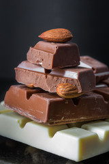 Pieces of milk chocolate with almonds and tiles of white chocolate with hazelnuts on a dark old glossy background