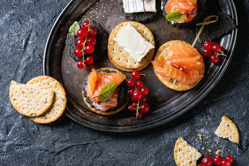 Stack of black charcoal and traditional crackers with smoked salmon, cream cheese, green salad and red currant berries on vintage metal tray over black stone background. Appetizer snack. Top view