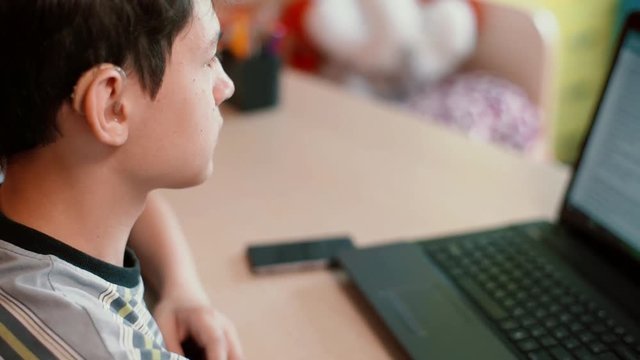 Hard of hearing teenager in hearing aids sitting at table using laptop