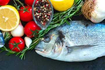Fresh raw dorado fish with rosemary, garlic, tomatoes, pepper and lemon on a black slate background. Selective focus. Overhead view. Copy space.