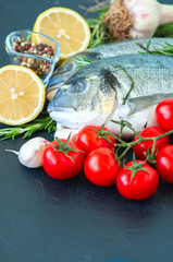 Fresh raw dorado fish with rosemary, garlic, tomatoes, pepper and lemon on a black slate background. Selective focus. Close up.