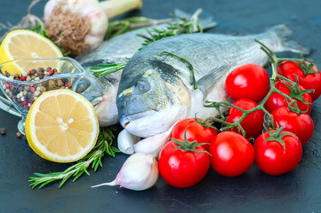 Fresh raw dorado fish with rosemary, garlic, tomatoes, pepper and lemon on a black slate background. Selective focus. Close up.