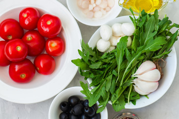 Ingredients for healthy salad. Tomatoes, rarugula, onion, mozzarella, olives, garlic and pepper with olive oil on a white stone background. Vegetarian food, health or cooking concept.