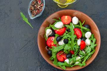 Close up of fresh salad with arugula, tomatoes, olives, onion, mozzarella and spices in a bowl on a gray slate background. Healthy food, Diet, Detox, Clean Eating or Vegetarian concept