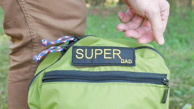 Men's hands cling to the Velcro of a green backpack word Super Dad. After the patch is hidden in a zip pocket