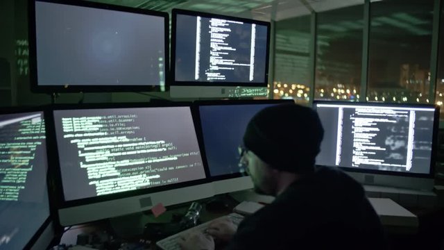 Tilt up of hacker sitting in the dark office and typing commands on several computer monitors