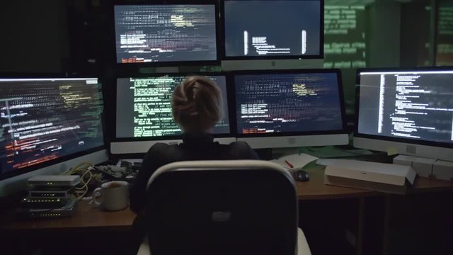 Rear view of female hacker sitting in the dark office in front of six monitors with data 