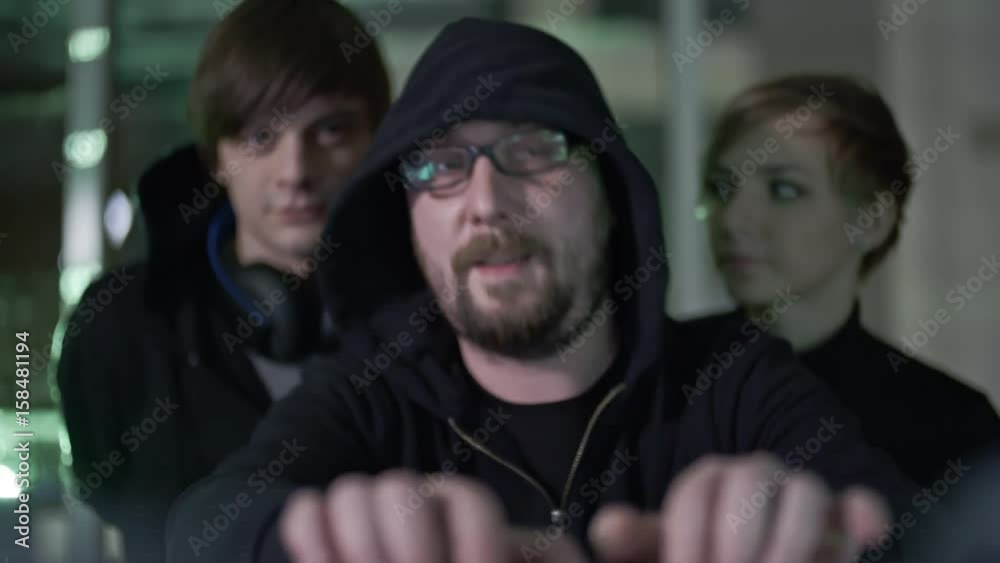 Wall mural bearded hacker in eyeglasses and hood working on computer with two teammates standing behind his bac