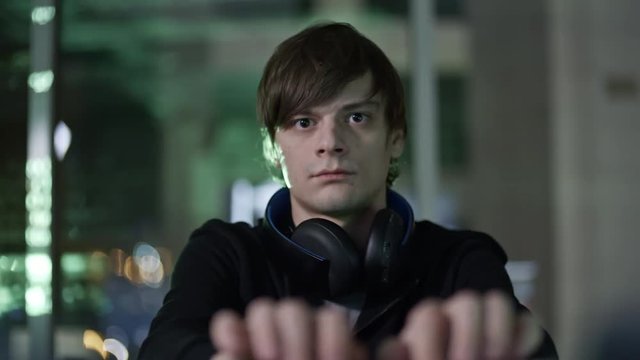 Concentrated young man with headphones around his neck typing on computer keyboard at desk in the dark room with green program codes reflected on walls