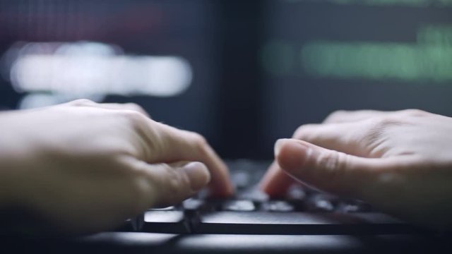 Tracking shot of hands of female hacker typing on computer keyboard; defocused screen with data in background