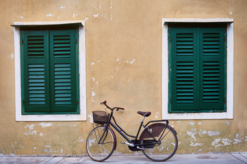 Fototapeta na wymiar Bicycle with metal basket parked in front of an old wall of a house with flaked yellow plaster and big widows with green shutters