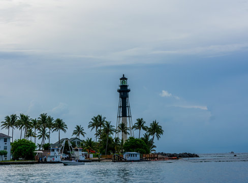 black and white lighthouse on a cloudy morning with boats and green palm trees along the rocky sandy shore