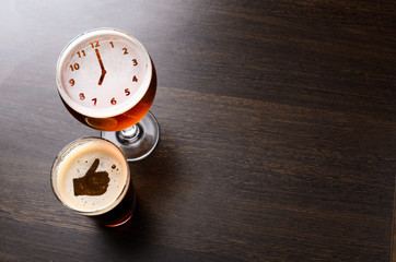 Thumbs up and clock silhouettes in two glasses of fresh beer on pub table, view from above