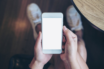 Mockup image of woman's hand holding white mobile phone with blank screen on thigh with wooden floor background in modern cafe