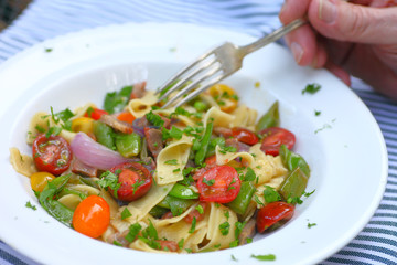 Bowl of noodles with cherry tomatoes and fresh peas with man’s hand and fork outdoors
