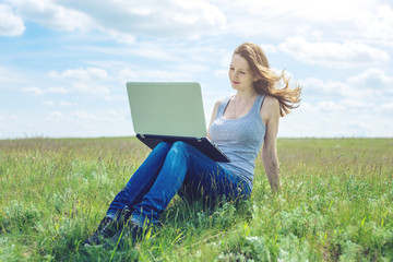 Woman sitting on a green meadow on the background of sky with clouds and working or studying with laptop wireless