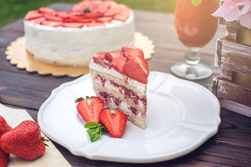 Delicious homemade summer cake with strawberries and butter cream on a wooden porch and a piece on the plate