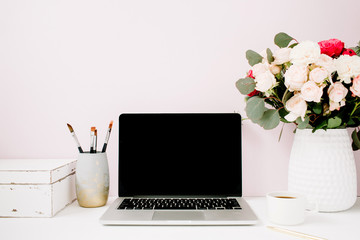 Home office desk with blank screen laptop, beautiful roses and eucalyptus bouquet, white vintage casket in front of pale pastel pink background. Blog, website or social media concept .