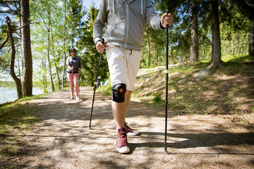 Man with bandage on knee. Rehabilitative sport in Finland - nordic walking. Man and mature woman hiking in green sunny forest. Active people outdoors. Scenic peaceful Finnish summer landscape.