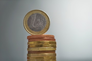 Side view of tall pile of metal coins in golden and copper color in front of silver background with one Euro (European currency EUR) coin on the top 