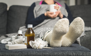 Cold medicine and sick woman drinking hot beverage to get well from flu, fever and virus. Dirty paper towels and tissues on table. Ill person wearing warm woolen stocking socks in winter.  - obrazy, fototapety, plakaty