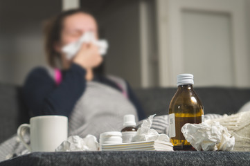 Sick woman sneezing to tissue. Medicine, hot beverage and dirty paper towels in front. Girl caught cold. Cough syrup and handkerchiefs on table. Very ill person feeling bad and having fever.