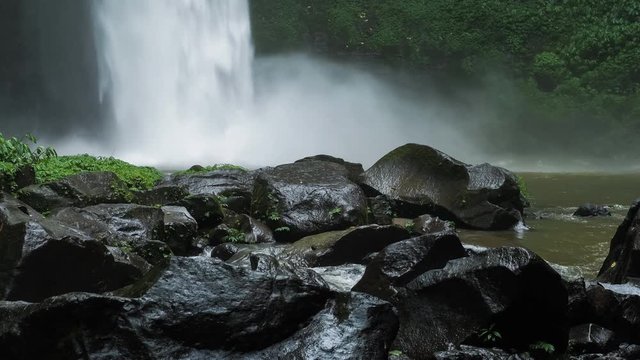 Close up of Nungnung waterfall, Falling water hitting water surface, some huge wet rocks in front of frame. Lush green leafes motion in background, Bali, Indonesia