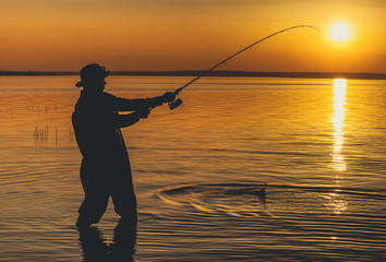 The fisherman catches fish for spinning and stands in the water against a beautiful sunset.