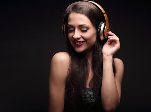 Beautiful laughing long hair young woman listening the music in wireless yellow headphone on dark black background. Closeup portrait.