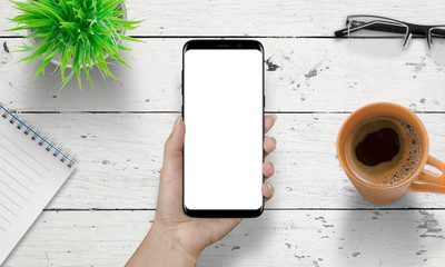 Smart phone in woman hand with isolated white screen for mockup. Wooden white desk in background with cup of coffee, plant, glasses beside. © Stanisic Vladimir