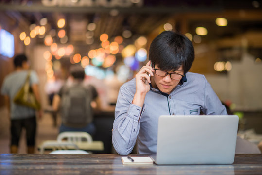 Young Asian Business Man Dressed In Casual Style Contact With Customer When Working In Cafe. Digital Nomad Lifestyle In Public Working Space.