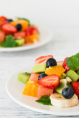 Healthy fresh fruit salad on white plate on wooden table. Close up. Delicious summer meal: strawberries, blueberries, oranges, kiwi, banana, watermelon, balm mint