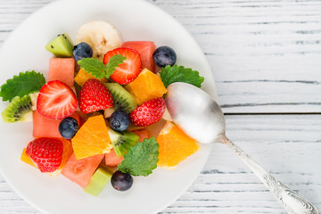 Healthy fresh fruit salad on white plate on wooden table. Top view. Delicious summer meal: strawberries, blueberries, oranges, kiwi, banana, watermelon, balm mint