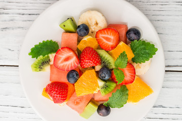 Healthy fresh fruit salad on white plate on wooden table. Top view. Delicious summer meal: strawberries, blueberries, oranges, kiwi, banana, watermelon, balm mint