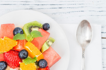 Healthy fresh fruit salad on white plate on wooden table. Top view. Delicious summer meal: strawberries, blueberries, oranges, kiwi, banana, watermelon, balm mint