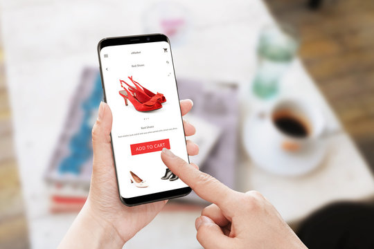 Add to cart red shoes on online store. Modern smart phone with round edges in woman hand. Table with coffee and magazines in background.