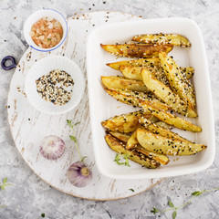 Slices of golden baked potato in black and white sesame seeds in a ceramic white background on a gray marble background with herbs and young garlic. Top View