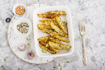 Slices of golden baked potato in black and white sesame seeds in a ceramic white background on a gray marble background with herbs and young garlic. Top View