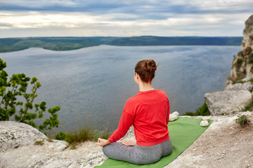 Fototapeta na wymiar Rear view of young woman sitting in lotus position on the rock over the river.