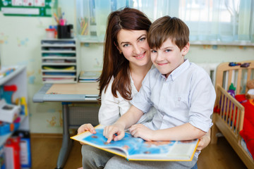 Beautiful happy mother with child boy 10 years old reading book, sitting on chair in children's room at home