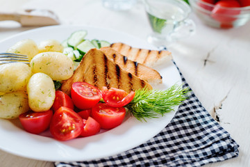Useful, hearty and delicious breakfast or dinner, boiled potato with dill, cherry tomato, cucumber, white bread toast, vegetable or olive oil and mineral sparkling water on a light wooden background