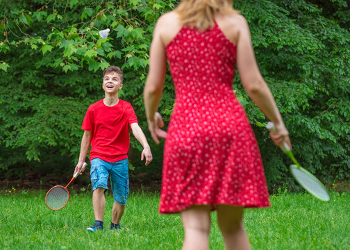 Mother And Son Playing Badminton In Meadow With Forest In Background. Child With Badminton Rackets In Hand. Kid Have Fun In Summer Park At Day.